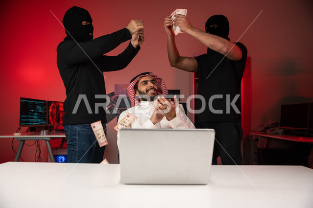 رجل عربي خليجي سعودي يجلس امام الحاسوب المحمول ، رجال ملثمين يستخدمون الاغراء المالي ، ابتزاز مالي ، الامن السيبراني ، الجرائم الالكترونية
