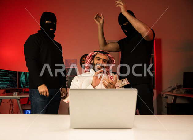 رجل عربي خليجي سعودي يجلس امام الحاسوب المحمول ، رجال ملثمين يستخدمون الاغراء المالي ، ابتزاز مالي ، الامن السيبراني ، الجرائم الالكترونية