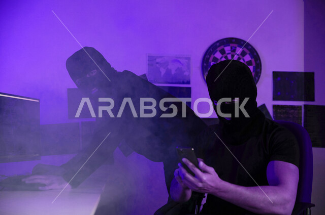 رجل ملثم يستخدم الهاتف المحمول ،  ملثم يجلس امام شاشات الحاسوب ، التجسس السيبراني ، الهجوم الالكتروني ، سرقة البيانات عبر الانترنت ، الجرائم الالكترونية ، الامن السيبراني