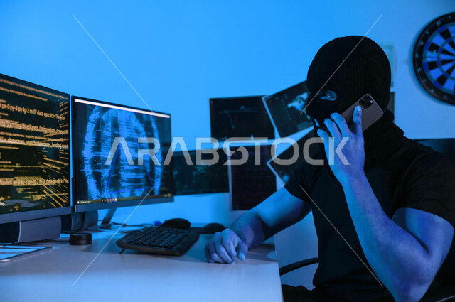 رجل ملثم يستخدم الهاتف المحمول ، يجلس امام شاشات الحاسوب ، التجسس السيبراني ، الهجوم الالكتروني ، سرقة البيانات عبر الانترنت ، الجرائم الالكترونية ، الامن السيبراني