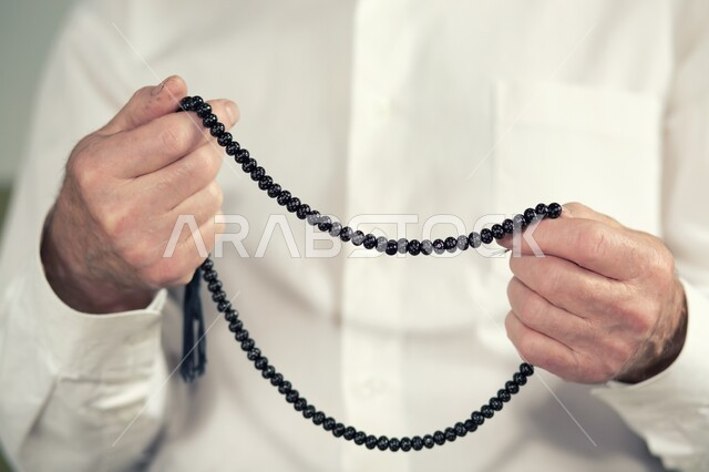 مفهوم العبادة والتقرب من الله تعالى, صورة مقربة لأيدي رجل عربي خليجي سعودي يرتدي الثوب التقليدي يحمل بيده سبحة سوداء يقوم بالتسبيح والدعاء, قراءة الأذكار والأدعية.