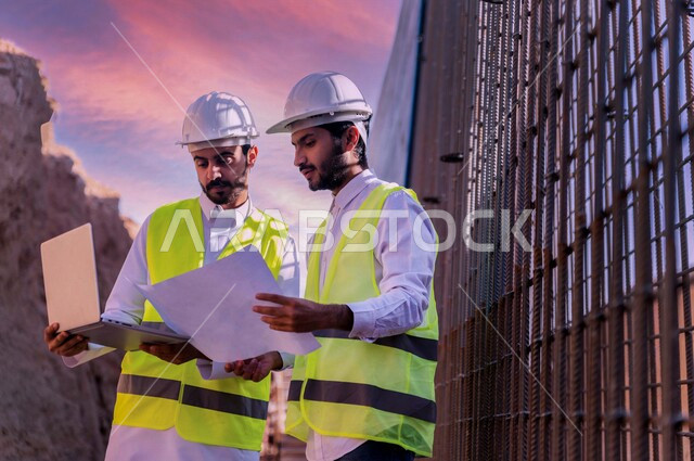 مقاول ومشرف العمل يرتديان خوذة وسترة خاصة بالعمل، عربيان خليجيان سعوديان يستخدمان الحاسوب المحمول وورقة مخطط البناء في موقع العمل ، مناقشة مخطط العمل ،اشراف ومتابعة العمل ، متابعة استراتيجية المشروع ، التصاميم المعمارية والانشائية