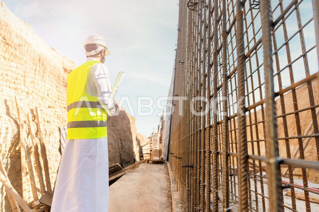 مشرف العمل عربي خليجي سعودي يرتدي خوذة وسترة خاصة بالعمل ، يمسك بيده ورقة مخطط البناء ، التصاميم المعمارية والانشائية ،الاشراف على العمل في موقع البناء ، متابعة استراتيجية المشروع