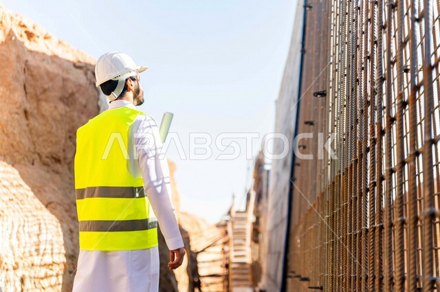 مشرف العمل عربي خليجي سعودي يرتدي خوذة وسترة خاصة بالعمل ، يمسك بيده ورقة مخطط البناء ، التصاميم المعمارية والانشائية ،الاشراف على العمل في موقع البناء ، متابعة استراتيجية المشروع