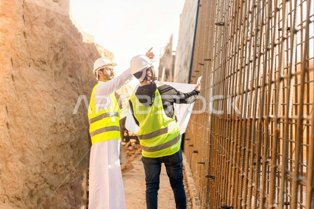 مقاول ومشرف العمل يرتديان خوذة وسترة خاصة بالعمل، عربيان خليجيان سعوديان يناقشان ورقة مخطط البناء ،اشراف ومتابعة العمل ، متابعة استراتيجية المشروع ، التصاميم المعمارية والانشائية