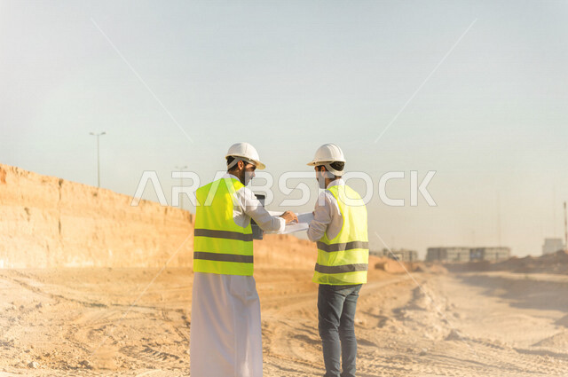 مقاول ومشرف العمل يرتديان خوذة وسترة خاصة بالعمل، عربيان خليجيان سعوديان يستخدمان الحاسوب المحمول وورقة مخطط البناء في موقع العمل ، مناقشة مخطط العمل ،اشراف ومتابعة العمل ، متابعة استراتيجية المشروع ، التصاميم المعمارية والانشائية