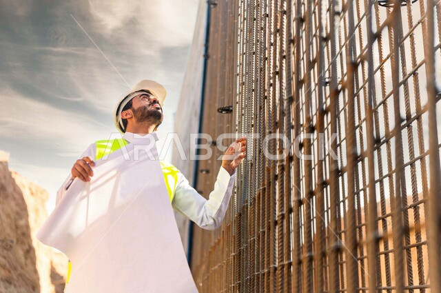 مشرف العمل عربي خليجي سعودي يرتدي خوذة وسترة خاصة بالعمل ، يمسك بيده ورقة مخطط البناء ، التصاميم المعمارية والانشائية ،الاشراف على العمل في موقع البناء ، متابعة استراتيجية المشروع