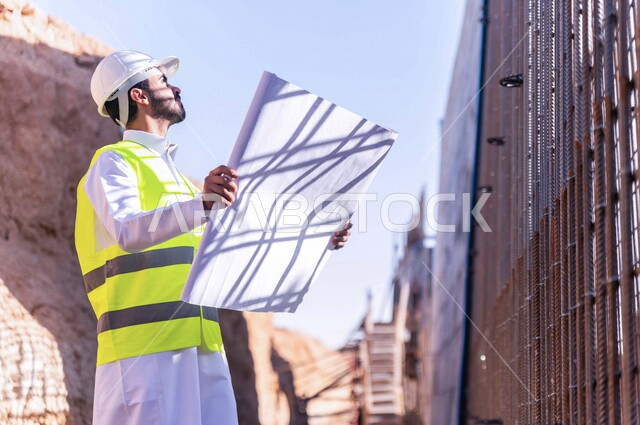 مشرف العمل عربي خليجي سعودي يرتدي خوذة وسترة خاصة بالعمل ، يمسك بيده ورقة مخطط البناء ، التصاميم المعمارية والانشائية ،الاشراف على العمل في موقع البناء ، متابعة استراتيجية المشروع