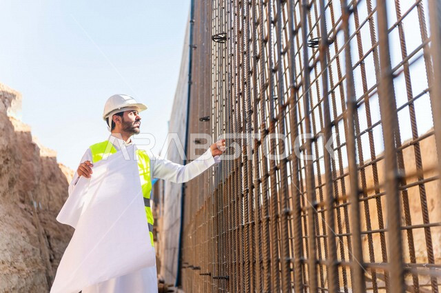 مشرف العمل عربي خليجي سعودي يرتدي خوذة وسترة خاصة بالعمل ، يمسك بيده ورقة مخطط البناء ، التصاميم المعمارية والانشائية ،الاشراف على العمل في موقع البناء ، متابعة استراتيجية المشروع
