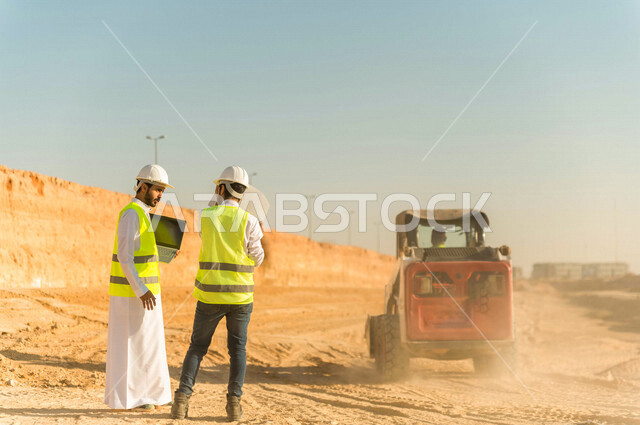 تصوير من الخلف لمقاول ومشرف العمل يرتديان خوذة وسترة خاصة بالعمل، عربيان خليجيان سعوديان يستخدمان الحاسوب المحمول في موقع العمل ، مناقشة العمل ،اشراف ومتابعة العمل ، متابعة استراتيجية المشروع ، التصاميم المعمارية والانشائية