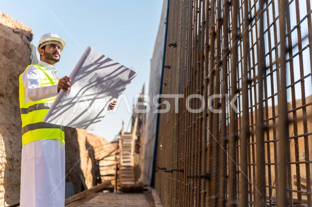 مشرف العمل عربي خليجي سعودي يرتدي خوذة وسترة خاصة بالعمل ، يمسك بيده ورقة مخطط البناء ، التصاميم المعمارية والانشائية ،الاشراف على العمل في موقع البناء ، متابعة استراتيجية المشروع