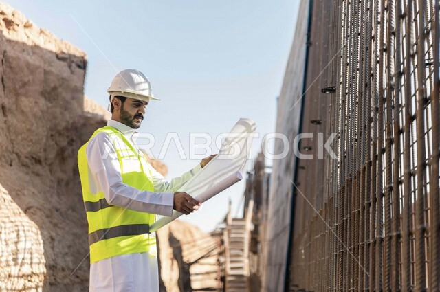 مشرف العمل عربي خليجي سعودي يرتدي خوذة وسترة خاصة بالعمل ، يمسك بيده ورقة مخطط البناء ، التصاميم المعمارية والانشائية ،الاشراف على العمل في موقع البناء ، متابعة استراتيجية المشروع
