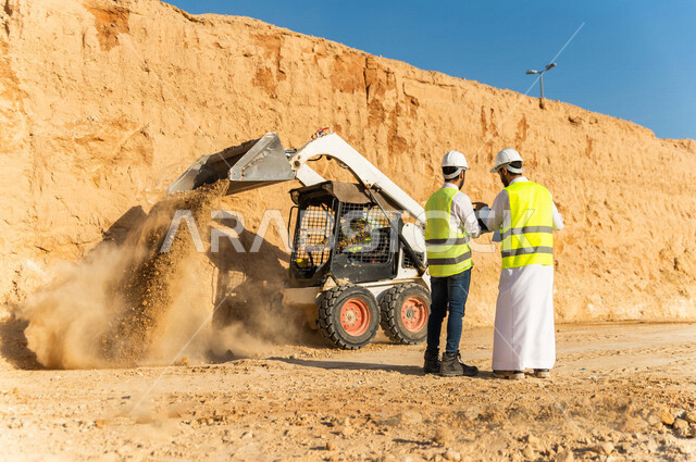 مقاول ومشرف العمل يرتديان خوذة وسترة خاصة بالعمل، عربيان خليجيان سعوديان يتناقشان في موقع العمل ،اشراف ومتابعة العمل ، متابعة استراتيجية المشروع ، التصاميم المعمارية والانشائية