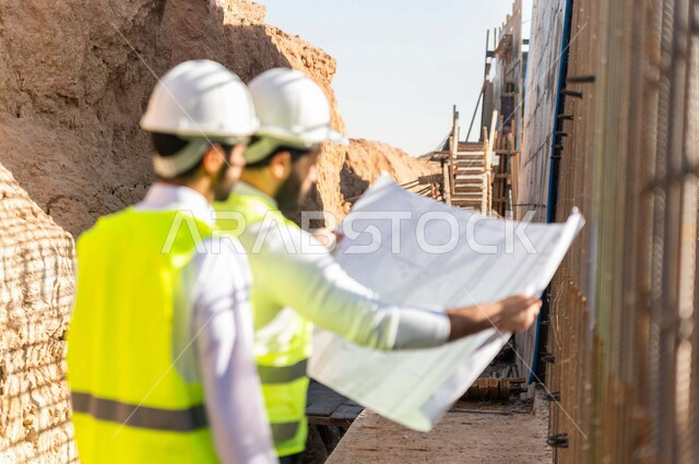 مقاول ومشرف العمل يرتديان خوذة وسترة خاصة بالعمل، عربيان خليجيان سعوديان يناقشان ورقة مخطط البناء ،اشراف ومتابعة العمل ، متابعة استراتيجية المشروع ، التصاميم المعمارية والانشائية