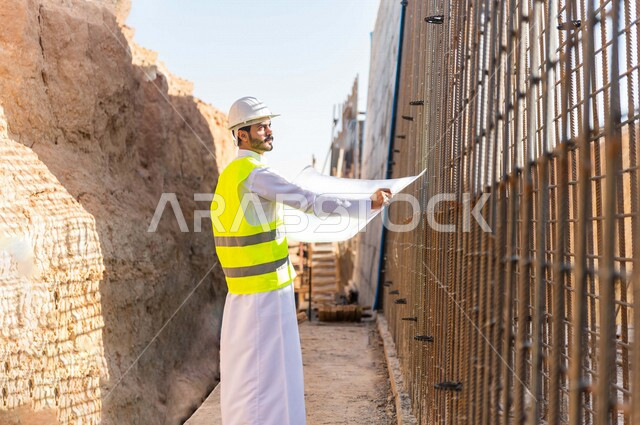 The work supervisor is an Arab, Gulf, Saudi, wearing a work helmet and jacket, holding in his hand a sheet of construction plan, architectural and construction designs, supervising work at the construction site, following up on the project strategy