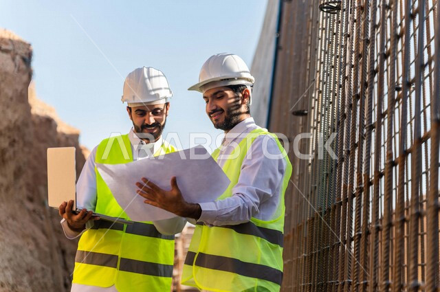 مقاول ومشرف العمل يرتديان خوذة وسترة خاصة بالعمل، عربيان خليجيان سعوديان يستخدمان الحاسوب المحمول وورقة مخطط البناء في موقع العمل ، مناقشة مخطط العمل ،اشراف ومتابعة العمل ، متابعة استراتيجية المشروع ، التصاميم المعمارية والانشائية