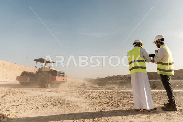 تصوير من الخلف لمقاول ومشرف العمل يرتديان خوذة وسترة خاصة بالعمل، عربيان خليجيان سعوديان يستخدمان الحاسوب المحمول في موقع العمل ، مناقشة العمل ،اشراف ومتابعة العمل ، متابعة استراتيجية المشروع ، التصاميم المعمارية والانشائية