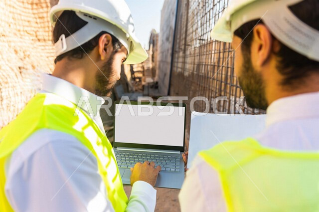 تصوير من الخلف لمقاول ومشرف العمل يرتديان خوذة وسترة خاصة بالعمل، عربيان خليجيان سعوديان يستخدمان الحاسوب المحمول في موقع العمل ، مناقشة العمل ،اشراف ومتابعة العمل ، متابعة استراتيجية المشروع ، التصاميم المعمارية والانشائية
