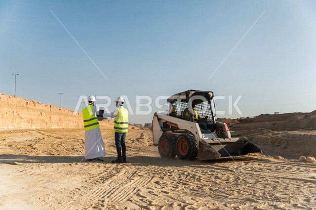 تصوير من الخلف لمقاول ومشرف العمل يرتديان خوذة وسترة خاصة بالعمل، عربيان خليجيان سعوديان يستخدمان الحاسوب المحمول في موقع العمل ، مناقشة العمل ،اشراف ومتابعة العمل ، متابعة استراتيجية المشروع ، التصاميم المعمارية والانشائية
