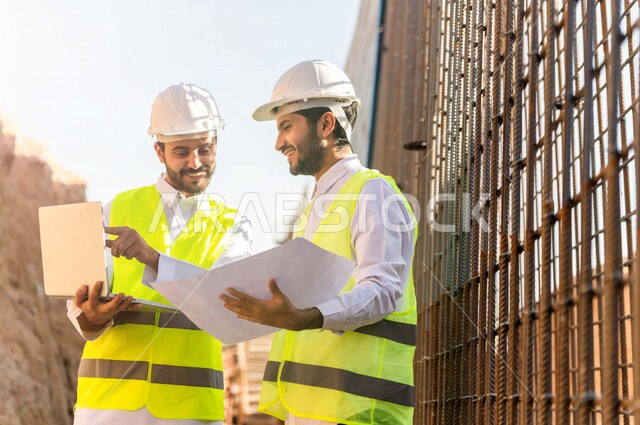 A contractor and a work supervisor wearing a work helmet and a work jacket, two Saudi Arabians using a laptop computer and a construction plan sheet at the work site, discussing the work plan, supervising and following up on work, following up on the proj