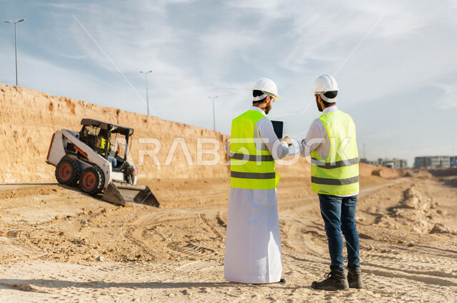 تصوير من الخلف لمقاول ومشرف العمل يرتديان خوذة وسترة خاصة بالعمل، عربيان خليجيان سعوديان يستخدمان الحاسوب المحمول في موقع العمل ، مناقشة العمل ،اشراف ومتابعة العمل ، متابعة استراتيجية المشروع ، التصاميم المعمارية والانشائية