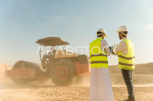 تصوير من الخلف لمقاول ومشرف العمل يرتديان خوذة وسترة خاصة بالعمل، عربيان خليجيان سعوديان يستخدمان الحاسوب المحمول في موقع العمل ، مناقشة العمل ،اشراف ومتابعة العمل ، متابعة استراتيجية المشروع ، التصاميم المعمارية والانشائية