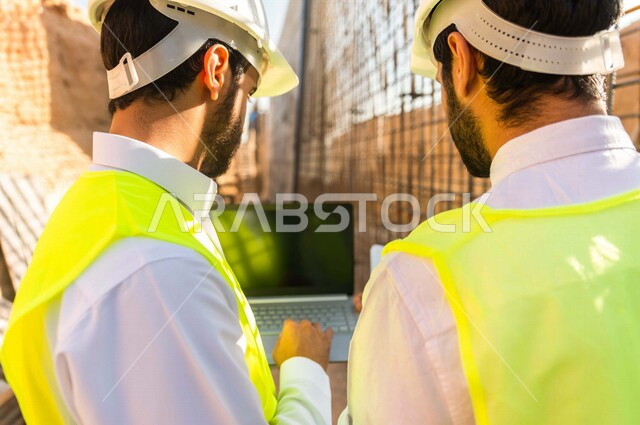 Photography from the back of a contractor and work supervisor wearing a work helmet and jacket, two Saudi Gulf Arabs using a laptop computer at the work site, discussing work, supervising and following up on work, following up on project strategy, archite