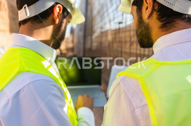 تصوير من الخلف لمقاول ومشرف العمل يرتديان خوذة وسترة خاصة بالعمل، عربيان خليجيان سعوديان يستخدمان الحاسوب المحمول في موقع العمل ، مناقشة العمل ،اشراف ومتابعة العمل ، متابعة استراتيجية المشروع ، التصاميم المعمارية والانشائية