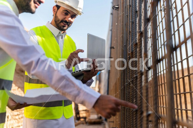 مقاول ومشرف العمل يرتديان خوذة وسترة خاصة بالعمل، عربيان خليجيان سعوديان يستخدمان الحاسوب المحمول وورقة مخطط البناء في موقع العمل ، مناقشة مخطط العمل ،اشراف ومتابعة العمل ، متابعة استراتيجية المشروع ، التصاميم المعمارية والانشائية