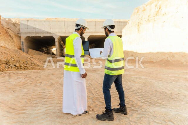مقاول ومشرف العمل يرتديان خوذة وسترة خاصة بالعمل، عربيان خليجيان سعوديان يستخدمان الحاسوب المحمول وورقة مخطط البناء في موقع العمل ، مناقشة مخطط العمل ،اشراف ومتابعة العمل ، متابعة استراتيجية المشروع ، التصاميم المعمارية والانشائية
