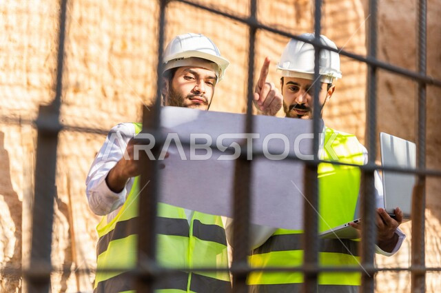 مقاول ومشرف العمل يرتديان خوذة وسترة خاصة بالعمل، عربيان خليجيان سعوديان يستخدمان الحاسوب المحمول وورقة مخطط البناء في موقع العمل ، مناقشة مخطط العمل ،اشراف ومتابعة العمل ، متابعة استراتيجية المشروع ، التصاميم المعمارية والانشائية