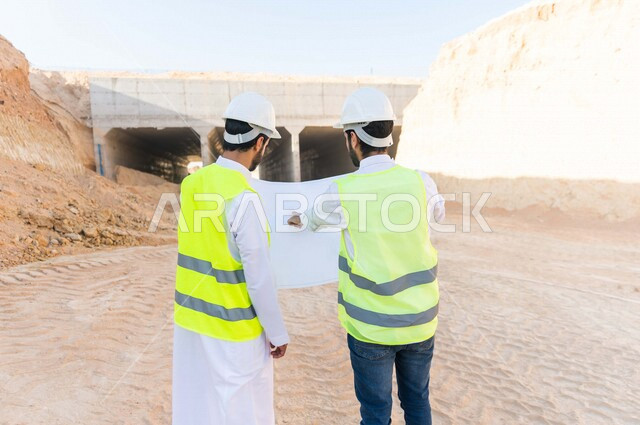 مقاول ومشرف العمل يرتديان خوذة وسترة خاصة بالعمل، عربيان خليجيان سعوديان يناقشان ورقة مخطط البناء ،اشراف ومتابعة العمل ، متابعة استراتيجية المشروع ، التصاميم المعمارية والانشائية