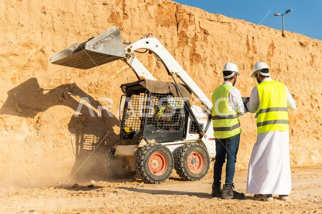 Photography from the back of a contractor and work supervisor wearing a work helmet and jacket, two Saudi Gulf Arabs using a laptop computer at the work site, discussing work, supervising and following up on work, following up on project strategy, archite