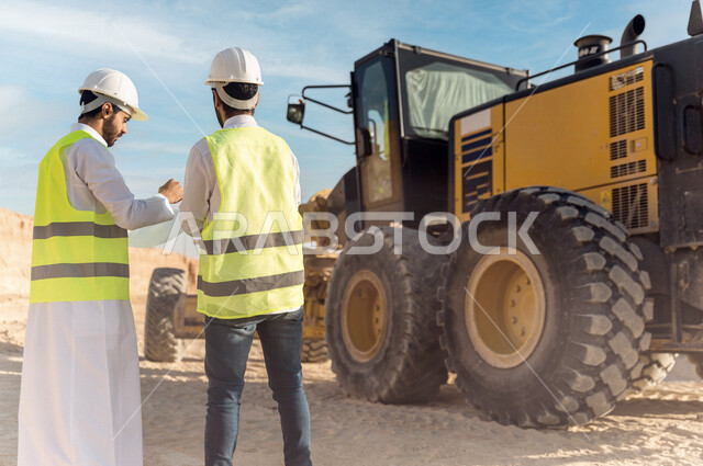 مقاول ومشرف العمل يرتديان خوذة وسترة خاصة بالعمل، عربيان خليجيان سعوديان يتناقشان في موقع العمل ،اشراف ومتابعة العمل ، متابعة استراتيجية المشروع ، التصاميم المعمارية والانشائية