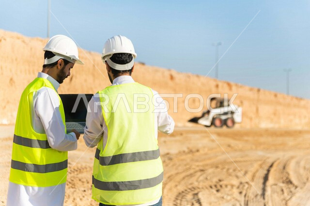 Photography from the back of a contractor and work supervisor wearing a work helmet and jacket, two Saudi Gulf Arabs using a laptop computer at the work site, discussing work, supervising and following up on work, following up on project strategy, archite