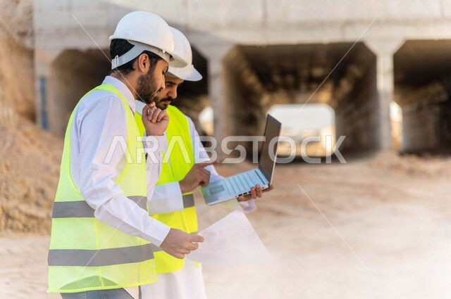 مقاول ومشرف العمل يرتديان خوذة وسترة خاصة بالعمل، عربيان خليجيان سعوديان يستخدمان الحاسوب المحمول وورقة مخطط البناء في موقع العمل ، مناقشة مخطط العمل ،اشراف ومتابعة العمل ، متابعة استراتيجية المشروع ، التصاميم المعمارية والانشائية