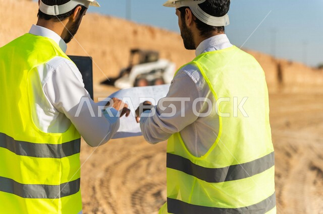 مقاول ومشرف العمل يرتديان خوذة وسترة خاصة بالعمل، عربيان خليجيان سعوديان يستخدمان الحاسوب المحمول وورقة مخطط البناء في موقع العمل ، مناقشة مخطط العمل ،اشراف ومتابعة العمل ، متابعة استراتيجية المشروع ، التصاميم المعمارية والانشائية
