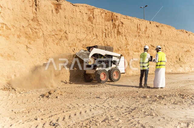 تصوير من الخلف لمقاول ومشرف العمل يرتديان خوذة وسترة خاصة بالعمل، عربيان خليجيان سعوديان يستخدمان الحاسوب المحمول في موقع العمل ، مناقشة العمل ،اشراف ومتابعة العمل ، متابعة استراتيجية المشروع ، التصاميم المعمارية والانشائية
