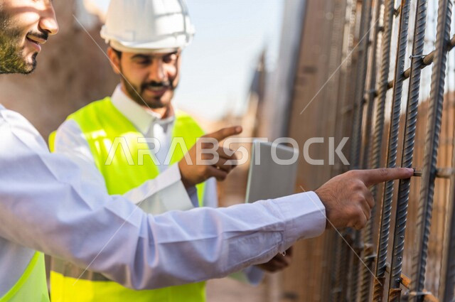 مقاول ومشرف العمل يرتديان خوذة وسترة خاصة بالعمل، عربيان خليجيان سعوديان يستخدمان الحاسوب المحمول في موقع العمل ، مناقشة العمل ،اشراف ومتابعة العمل ، متابعة استراتيجية المشروع ، التصاميم المعمارية والانشائية