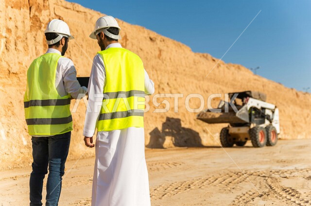 تصوير من الخلف لمقاول ومشرف العمل يرتديان خوذة وسترة خاصة بالعمل، عربيان خليجيان سعوديان يستخدمان الحاسوب المحمول في موقع العمل ، مناقشة العمل ،اشراف ومتابعة العمل ، متابعة استراتيجية المشروع ، التصاميم المعمارية والانشائية