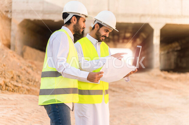 مقاول ومشرف العمل يرتديان خوذة وسترة خاصة بالعمل، عربيان خليجيان سعوديان يستخدمان الحاسوب المحمول وورقة مخطط البناء في موقع العمل ، مناقشة مخطط العمل ،اشراف ومتابعة العمل ، متابعة استراتيجية المشروع ، التصاميم المعمارية والانشائية
