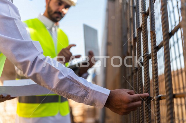 A contractor and a work supervisor wearing a work helmet and a work jacket, two Saudi Arabians using a laptop computer and a construction plan sheet at the work site, discussing the work plan, supervising and following up on work, following up on the proj