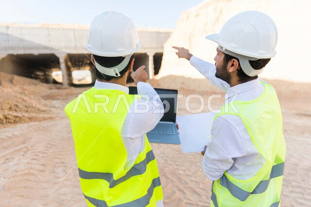 مقاول ومشرف العمل يرتديان خوذة وسترة خاصة بالعمل، عربيان خليجيان سعوديان يستخدمان الحاسوب المحمول وورقة مخطط البناء في موقع العمل ، مناقشة مخطط العمل ،اشراف ومتابعة العمل ، متابعة استراتيجية المشروع ، التصاميم المعمارية والانشائية