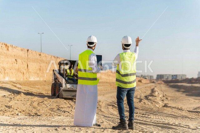 تصوير من الخلف لمقاول ومشرف العمل يرتديان خوذة وسترة خاصة بالعمل، عربيان خليجيان سعوديان يستخدمان الحاسوب المحمول في موقع العمل ، مناقشة العمل ،اشراف ومتابعة العمل ، متابعة استراتيجية المشروع ، التصاميم المعمارية والانشائية