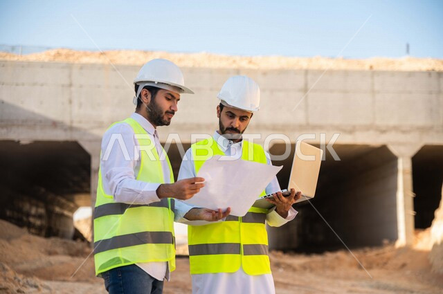 مقاول ومشرف العمل يرتديان خوذة وسترة خاصة بالعمل، عربيان خليجيان سعوديان يستخدمان الحاسوب المحمول وورقة مخطط البناء في موقع العمل ، مناقشة مخطط العمل ،اشراف ومتابعة العمل ، متابعة استراتيجية المشروع ، التصاميم المعمارية والانشائية