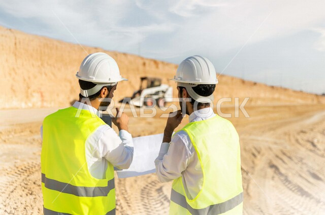 مقاول ومشرف العمل يرتديان خوذة وسترة خاصة بالعمل، عربيان خليجيان سعوديان يناقشان ورقة مخطط البناء ،اشراف ومتابعة العمل ، متابعة استراتيجية المشروع ، التصاميم المعمارية والانشائية