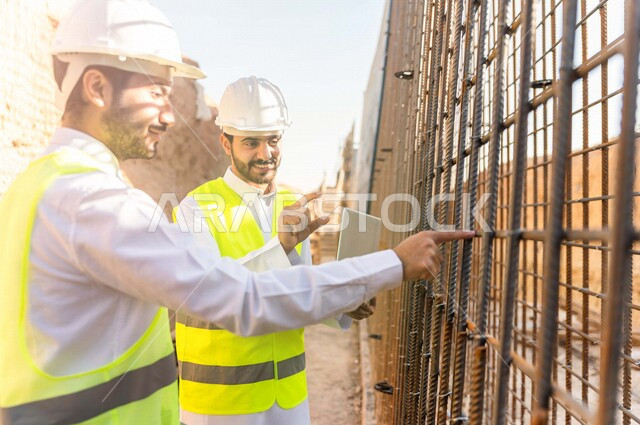 مقاول ومشرف العمل يرتديان خوذة وسترة خاصة بالعمل، عربيان خليجيان سعوديان يستخدمان الحاسوب المحمول في موقع العمل ، مناقشة العمل ،اشراف ومتابعة العمل ، متابعة استراتيجية المشروع ، التصاميم المعمارية والانشائية
