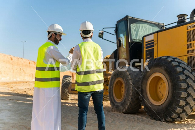مقاول ومشرف العمل يرتديان خوذة وسترة خاصة بالعمل، عربيان خليجيان سعوديان يتناقشان في موقع العمل ،اشراف ومتابعة العمل ، متابعة استراتيجية المشروع ، التصاميم المعمارية والانشائية