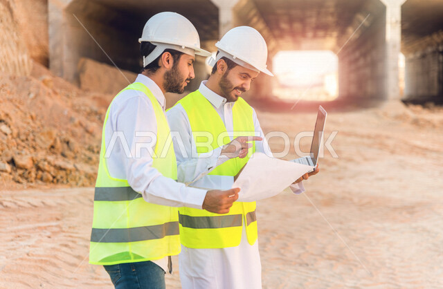 مقاول ومشرف العمل يرتديان خوذة وسترة خاصة بالعمل، عربيان خليجيان سعوديان يستخدمان الحاسوب المحمول وورقة مخطط البناء في موقع العمل ، مناقشة مخطط العمل ،اشراف ومتابعة العمل ، متابعة استراتيجية المشروع ، التصاميم المعمارية والانشائية