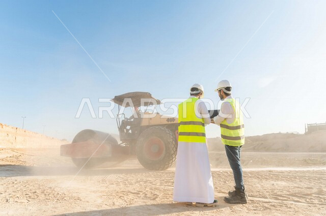 تصوير من الخلف لمقاول ومشرف العمل يرتديان خوذة وسترة خاصة بالعمل، عربيان خليجيان سعوديان يستخدمان الحاسوب المحمول في موقع العمل ، مناقشة العمل ،اشراف ومتابعة العمل ، متابعة استراتيجية المشروع ، التصاميم المعمارية والانشائية